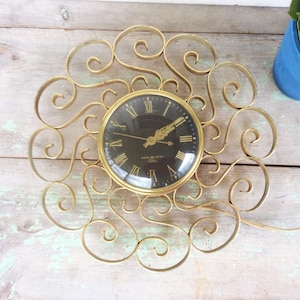 以下が含まれることがあります： 黒い文字盤と金色のローマ数字が特徴の、ヴィンテージのゼネラル・エレクトリックの壁掛け時計。時計は金色の装飾的で渦巻く金属フレームに収められています。時計の針は金色で、電気時計です。