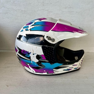 May include: White, black, and multi-colored motocross helmet with a visor. The helmet features a design with blue, purple, and black accents. The helmet has some wear and tear.