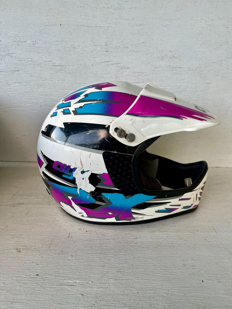 May include: White, black, and multi-colored motocross helmet with a visor. The helmet features a design with blue, purple, and black accents. The helmet has some wear and tear.