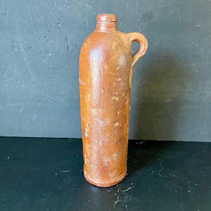 Puede incluir: Una botella de gres marrón con asa. La botella tiene un fondo redondeado y un cuello ligeramente acampanado.