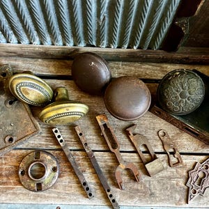 May include: An assortment of antique door hardware, including doorknobs, plates, and various metal pieces. The doorknobs are in shades of brown and gold, with intricate designs. The hardware appears aged and weathered, suggesting a vintage aesthetic.