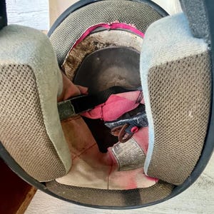 May include: An open view of the inside of a black motorcycle helmet. The interior features a pink and white lining, with tan mesh padding on either side. A black strap and a tag are visible.
