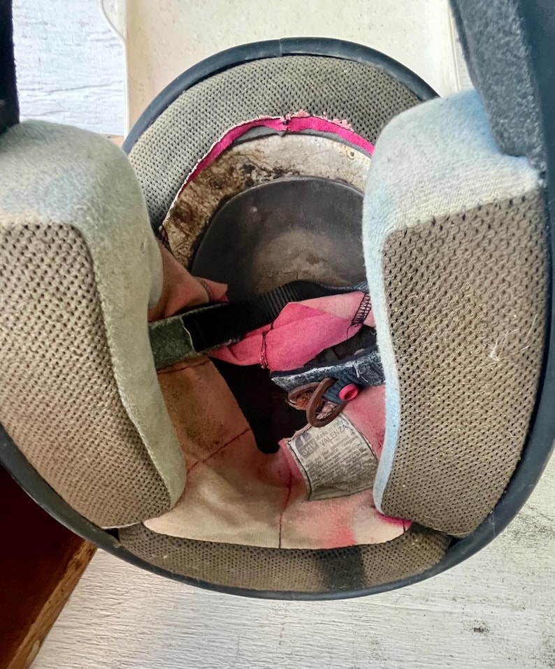 May include: An open view of the inside of a black motorcycle helmet. The interior features a pink and white lining, with tan mesh padding on either side. A black strap and a tag are visible.
