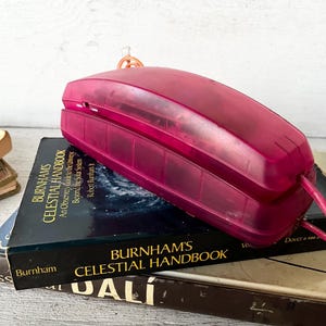 May include: A translucent magenta plastic landline telephone handset resting on top of a stack of books. The top book is titled Burnham's Celestial Handbook, An Observer's Guide to the Universe, Beyond the Solar System by Robert Burnham Jr.