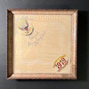 May include: A framed embroidered artwork featuring an eagle emblem and text. The frame is a light brown with a decorative edge. The artwork includes the text "Edward Young England" and a patch that says "Souvenir de France 1919".