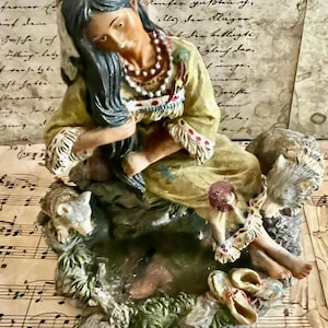 May include: A detailed figurine of a Native American woman in a yellow dress, adorned with a beaded necklace and feather. The sculpture includes a wolf and a pair of moccasins, set on a base with greenery and musical notes.