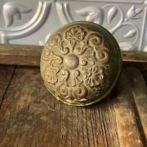 May include: Antique bronze door knob with an ornate floral design. The knob has a round shape and a weathered, aged appearance. The floral pattern is centered on the knob's surface.