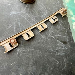 May include: A silver metal car emblem with the word "DODGE" embossed in raised letters. The emblem has two small screws on the back.
