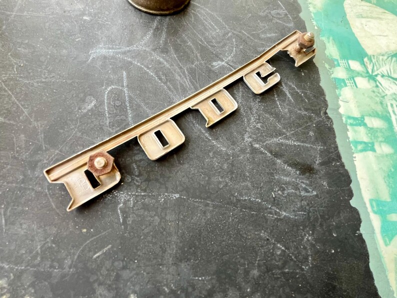 May include: A silver metal car emblem with the word "DODGE" embossed in raised letters. The emblem has two small screws on the back.