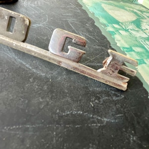May include: A vintage silver-colored Dodge car emblem with the word "Dodge" in raised letters.