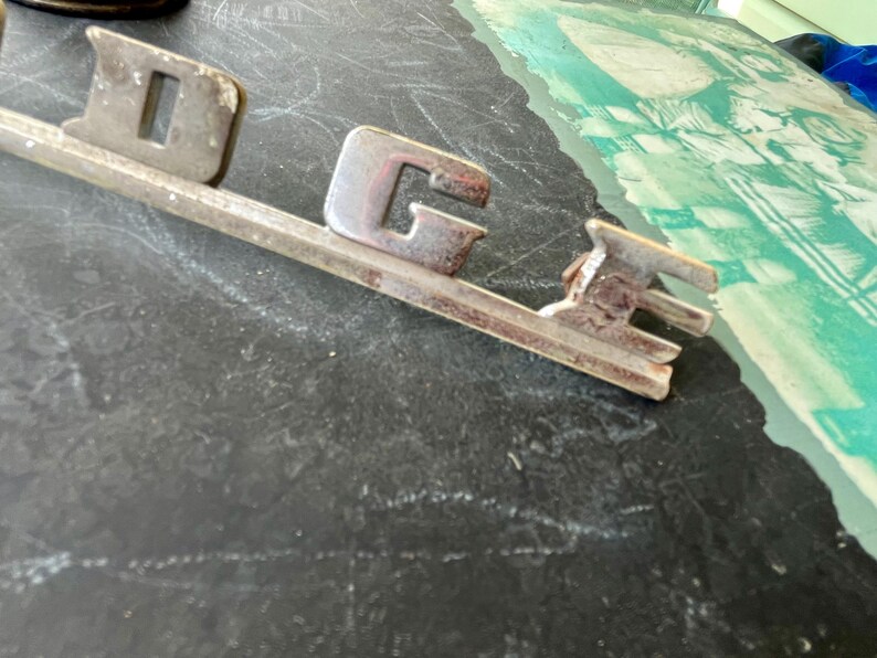 May include: A vintage silver-colored Dodge car emblem with the word "Dodge" in raised letters.