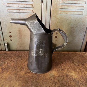 May include: Vintage metal oil can with a spout and handle. The can is dark gray with the text "ATLANTIC 2 Oz. Ltd. U.S.A. T.D." embossed on the side. The can has a spout and handle and is sitting on a rusty metal surface.