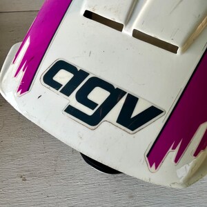 May include: A white and purple motorcycle helmet with the brand name "AGV" in black lettering. The helmet has a few vents and some scratches. The purple accents are on the sides and top of the helmet.