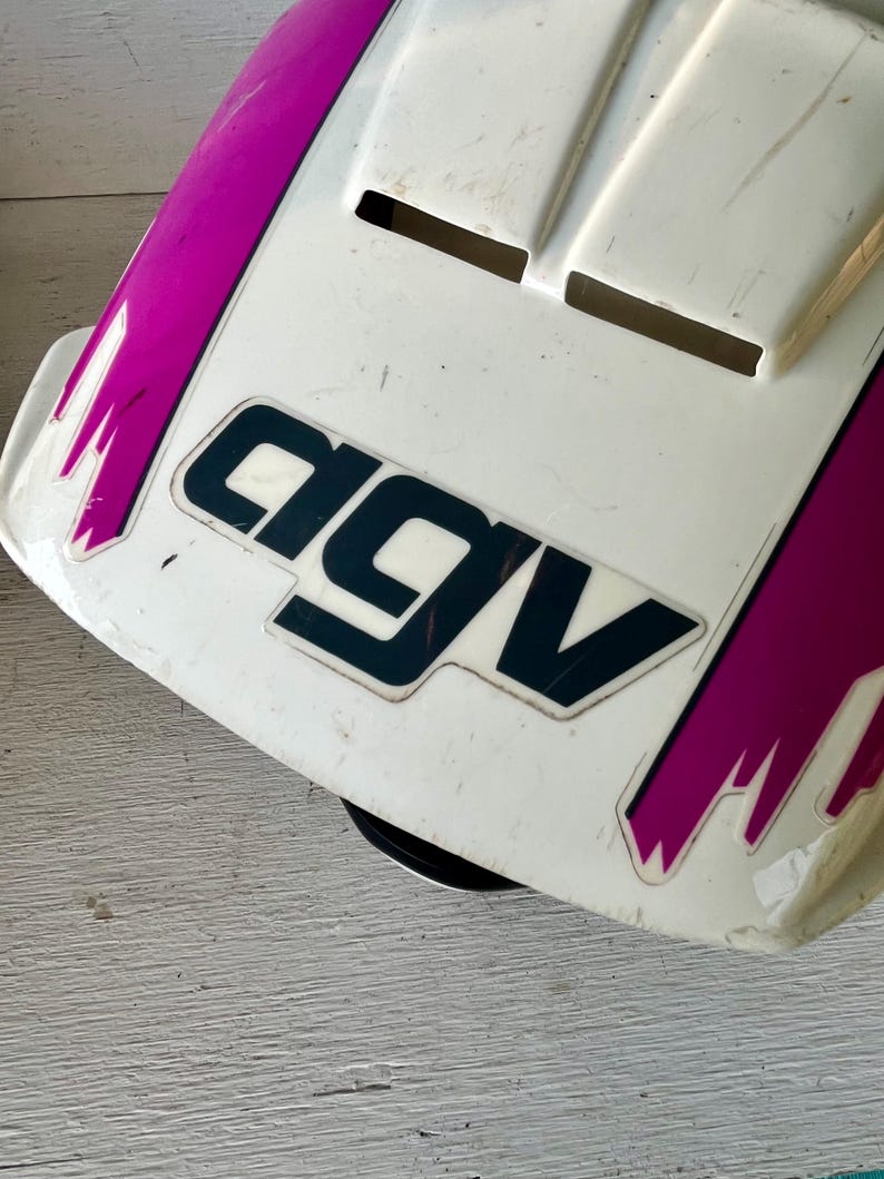May include: A white and purple motorcycle helmet with the brand name "AGV" in black lettering. The helmet has a few vents and some scratches. The purple accents are on the sides and top of the helmet.