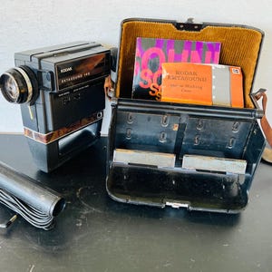 May include: A vintage Kodak Ektasound 140 movie camera with a black body and a brown wood-grain accent. The camera is next to a black case with a brown interior, containing books and accessories. A black microphone is also present.