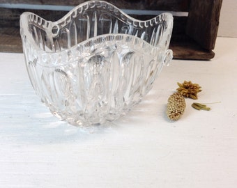 May include: A clear, decorative glass bowl with a scalloped rim and intricate cut-glass detailing. The bowl has small handles on each side. Dried floral elements are next to the bowl on a white surface.