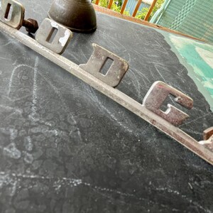 May include: A silver metal car emblem with the word "DODGE" spelled out in raised letters.