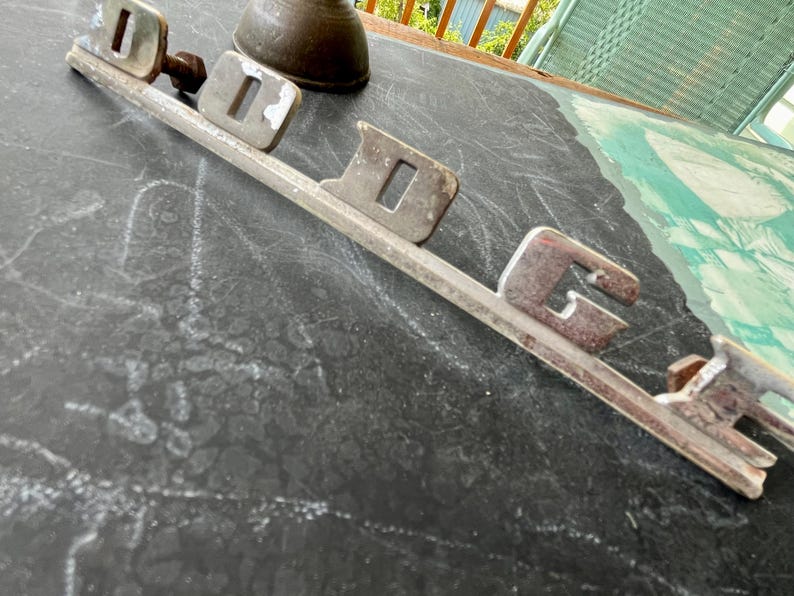May include: A silver metal car emblem with the word "DODGE" spelled out in raised letters.