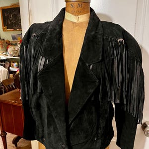 May include: Black suede jacket with fringe detailing on the sleeves. The jacket has a notched collar, button closures, and a tailored fit. The jacket is displayed on a vintage dress form.