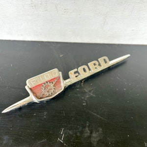 May include: A vintage Ford F-100 emblem, with the word "FORD" in silver lettering. The emblem has a red and silver rectangular section with the number "F-100". The emblem is on a black surface.