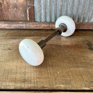 May include: Antique white porcelain door knob with a dark metal stem. The knob has an oval shape and is set against a rustic wooden background. A vintage hardware piece.
