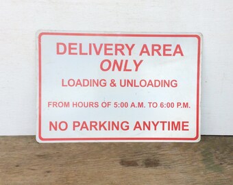Puede incluir: Un letrero rectangular de metal con fondo blanco y borde rojo. El letrero dice "DELIVERY AREA ONLY" y "LOADING & UNLOADING" con horarios de funcionamiento. También se muestra "NO PARKING ANYTIME".