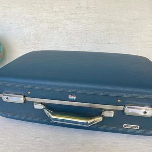 Maleta rígida vintage AMERICAN TOURISTER azul con 2 cierres de metal plateados y borde, interior de tela satinada con diseño floral azul claro y divisor
