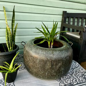 May include: A large, weathered metal planter with two handles, holding a green plant. Two smaller black pots with plants are to the left. The scene is set on a patterned rug, with a wooden bench in the background.