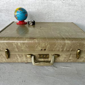 May include: A rectangular beige suitcase with a marbled pattern, featuring gold-toned metal latches and a central locking handle. Resting on top are a small, colorful globe on a red base and a miniature black camera with a silver lens detail.