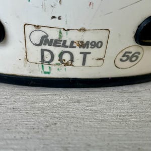 May include: Close-up of a white helmet with visible wear and tear. The helmet features a sticker with the text "Snell M90 DOT" and a size marking of "56". The helmet has black accents and is designed for protection.