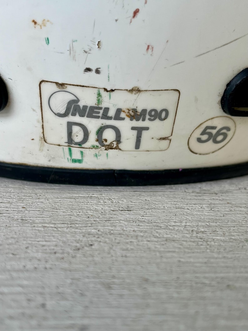 May include: Close-up of a white helmet with visible wear and tear. The helmet features a sticker with the text "Snell M90 DOT" and a size marking of "56". The helmet has black accents and is designed for protection.