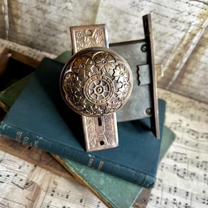 May include: Antique brass door knob and lock set with floral detailing. The round knob features an intricate flower design. The set is placed on top of a stack of vintage books with green covers, creating a classic, vintage aesthetic.