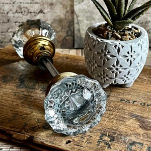 May include: Clear glass doorknob with a faceted design and brass hardware. The doorknob is lying on a weathered wooden surface. A small, gray decorative pot with a succulent plant is in the background.