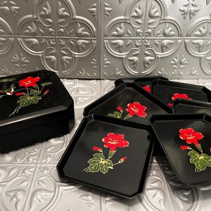 May include: A set of black lacquerware consisting of a lidded box and five matching square plates. Each piece features a hand-painted design of vibrant red flowers with green leaves. The lidded box also includes a small hummingbird illustration.