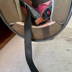May include: Interior view of a used black motorcycle helmet. The inside features brown mesh padding, a black chin strap with a red snap, and a pink fabric lining. The helmet shows signs of wear.