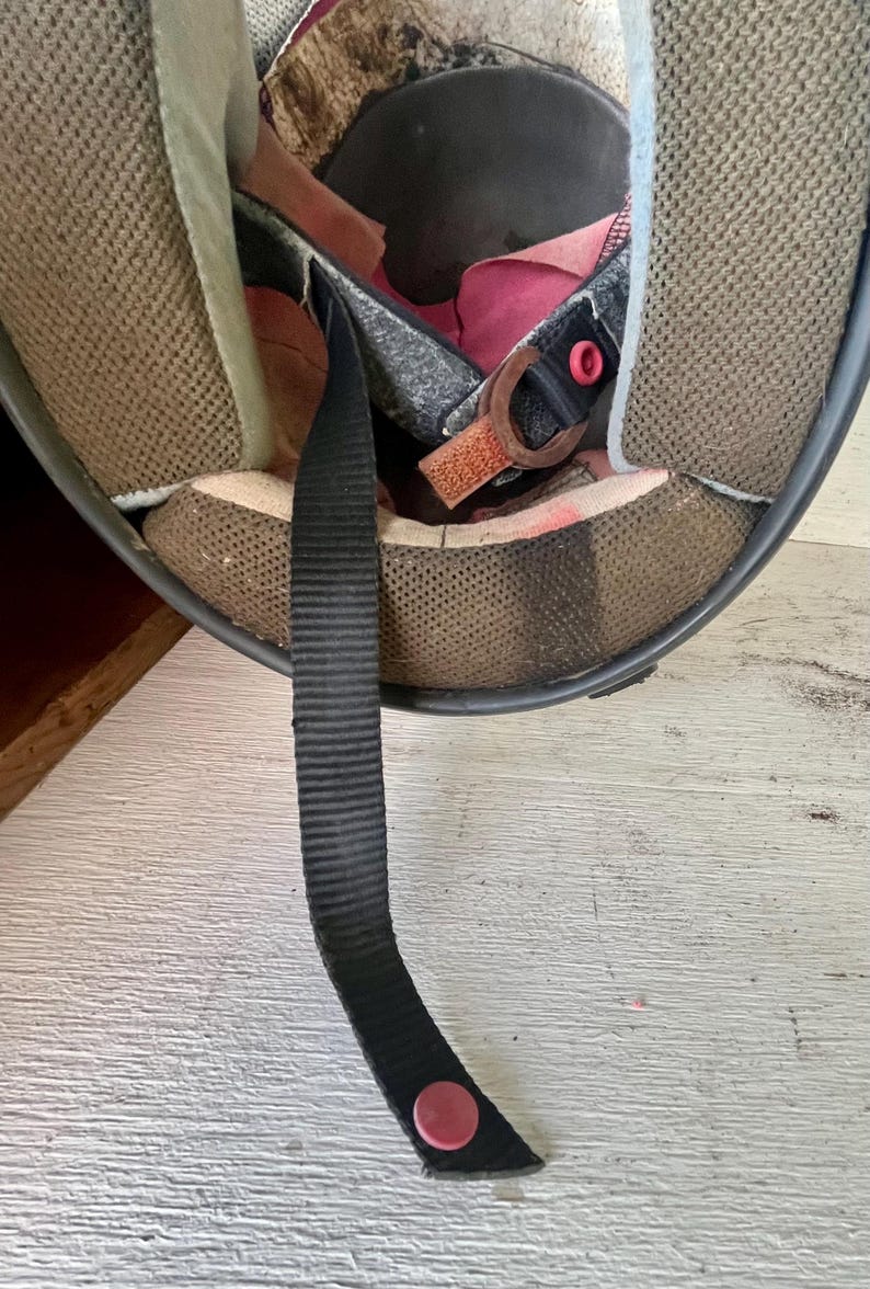 May include: Interior view of a used black motorcycle helmet. The inside features brown mesh padding, a black chin strap with a red snap, and a pink fabric lining. The helmet shows signs of wear.
