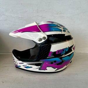 May include: White, black, and multi-colored motorcycle helmet with a visor. The helmet features a design with purple, blue, and white geometric shapes. The text "AGV" is visible on the side of the helmet.