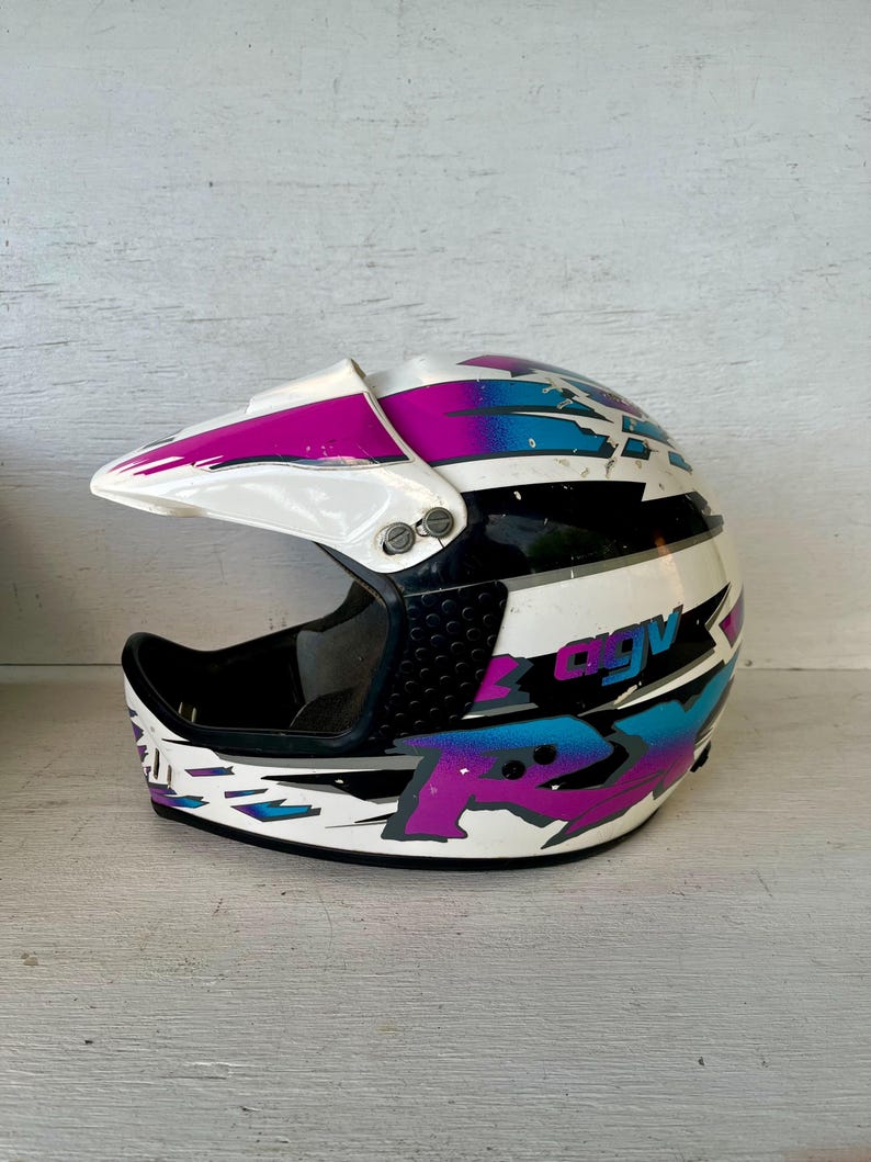 May include: White, black, and multi-colored motorcycle helmet with a visor. The helmet features a design with purple, blue, and white geometric shapes. The text "AGV" is visible on the side of the helmet.