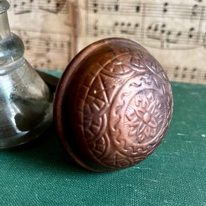 May include: A close-up of a vintage, copper-colored door knob with intricate floral and geometric patterns. The knob is round and has a warm, antique finish. A clear glass bottle is partially visible in the background.
