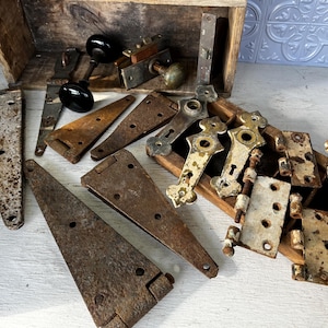 May include: A collection of vintage hardware components, including hinges, doorknobs, and latches, displayed on a wooden surface. The metal pieces exhibit a weathered, rusty patina, with varying shapes and sizes. Some hinges are triangular, while others are rectangular, and the doorknobs are black.