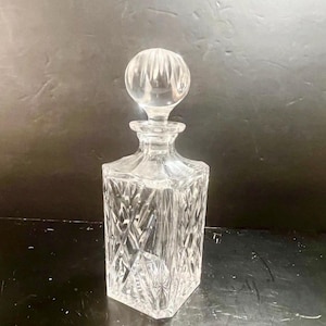 May include: A clear, square-shaped glass decanter with a round stopper. The decanter features a diamond-cut pattern on the body and a spherical stopper with vertical grooves. The design is classic and elegant, suitable for serving spirits.