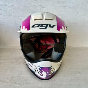 May include: White and pink AGV motorcycle helmet with black interior padding. The helmet has a white base with pink and purple accents. The AGV logo is prominently displayed in black. The helmet is designed for safety and protection.