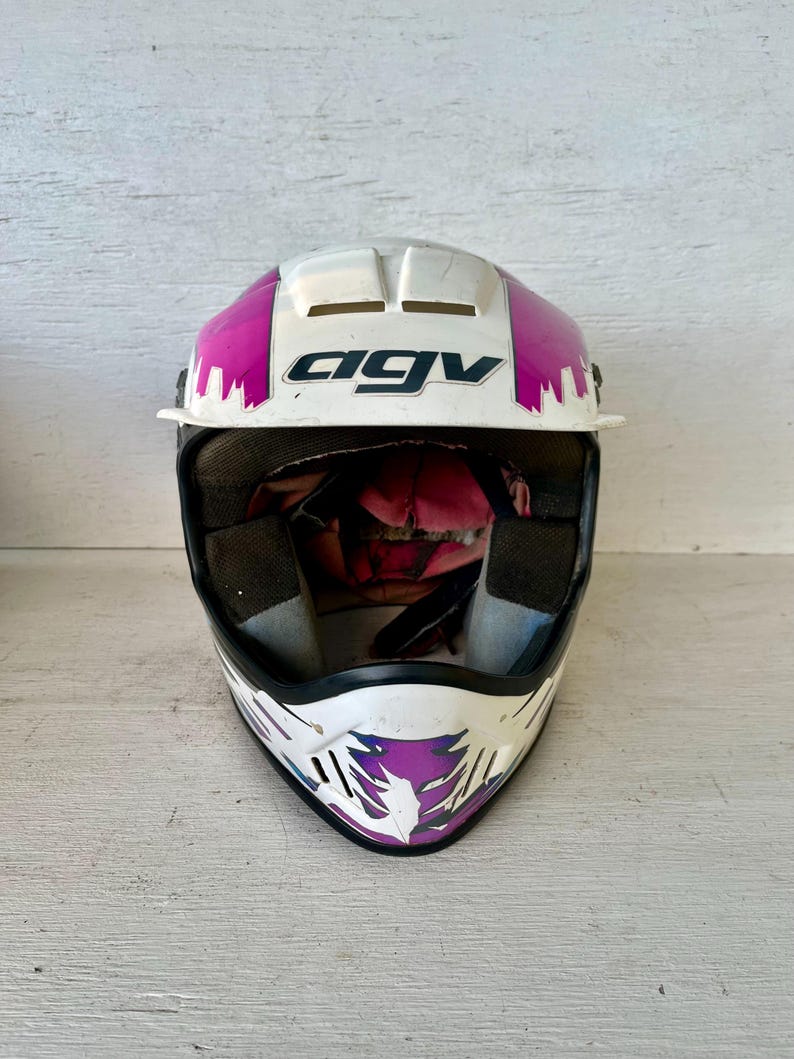 May include: White and pink AGV motorcycle helmet with black interior padding. The helmet has a white base with pink and purple accents. The AGV logo is prominently displayed in black. The helmet is designed for safety and protection.