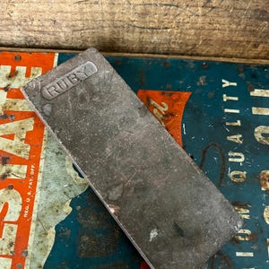 May include: A rectangular, weathered metal sharpening stone with the word "RUBY" embossed on one end. The stone is gray and shows signs of wear. It rests on a vintage sign with red, white, and blue colors.