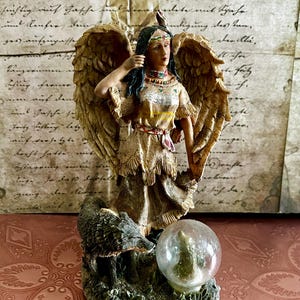 May include: A detailed figurine of an angel with feathered wings, wearing a Native American-inspired dress and headdress. The angel stands above a wolf and a snow globe. The color palette includes beige, brown, and green.