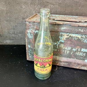May include: A vintage glass bottle of Royal Crown Cola. The bottle is clear and has a textured surface. The label is red, yellow, and black with the text "Royal Crown Cola" and "RC Cola". The bottle is sitting on top of a rusty metal box.