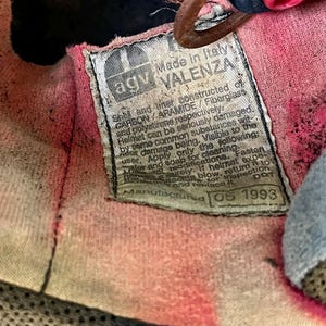 May include: Close-up of the interior of a vintage AGV Valenza helmet. The label reads "Made in Italy" and includes manufacturing details. The interior fabric is pink and shows signs of wear.