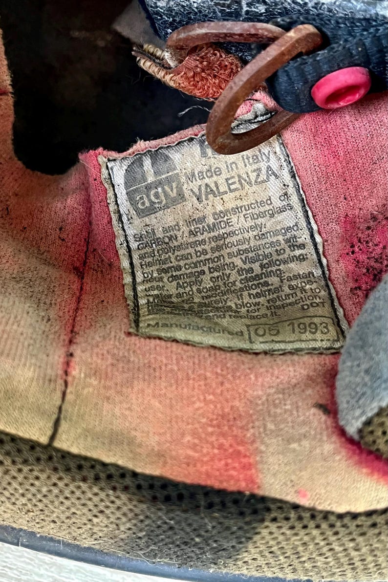 May include: Close-up of the interior of a vintage AGV Valenza helmet. The label reads "Made in Italy" and includes manufacturing details. The interior fabric is pink and shows signs of wear.