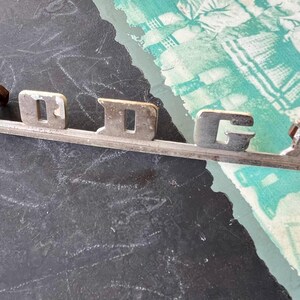 May include: A silver metal emblem with the word "DODGE" in raised letters. The emblem is on a black surface.