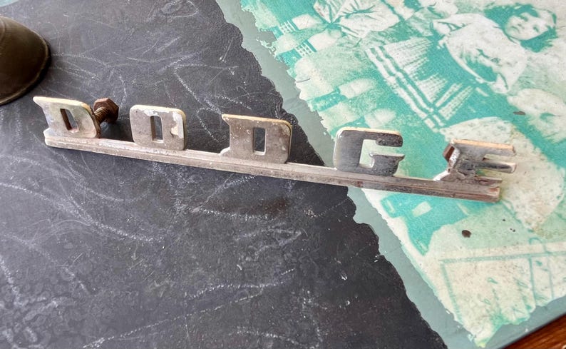 May include: A silver metal emblem with the word "DODGE" in raised letters. The emblem is on a black surface.